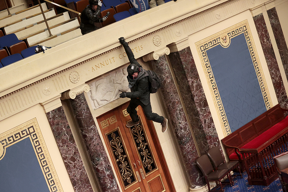 A photograph of a black-clad rioter hanging off of the press gallery of the House chambers.